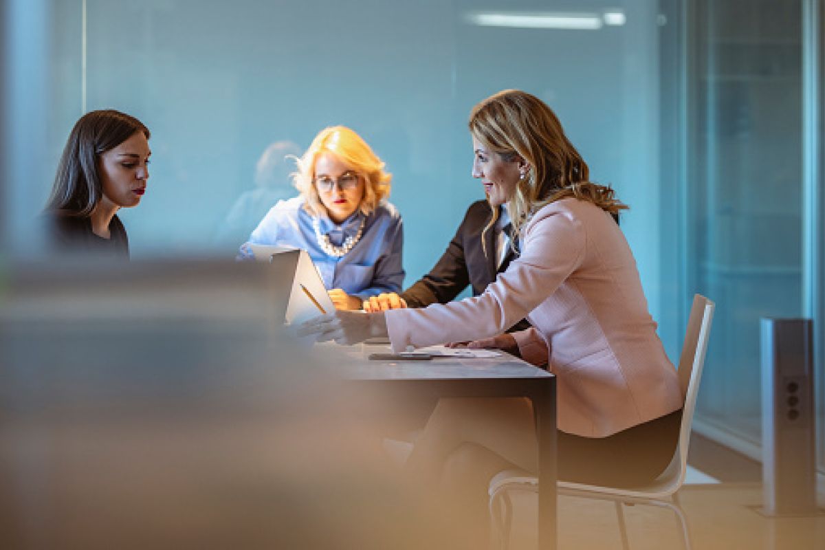 A group of female business leaders from various backgrounds strategize over a project in a well-lit, contemporary office space, utilizing digital tools to enhance productivity.