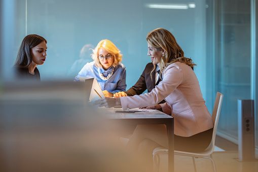 A group of female business leaders from various backgrounds strategize over a project in a well-lit, contemporary office space, utilizing digital tools to enhance productivity.