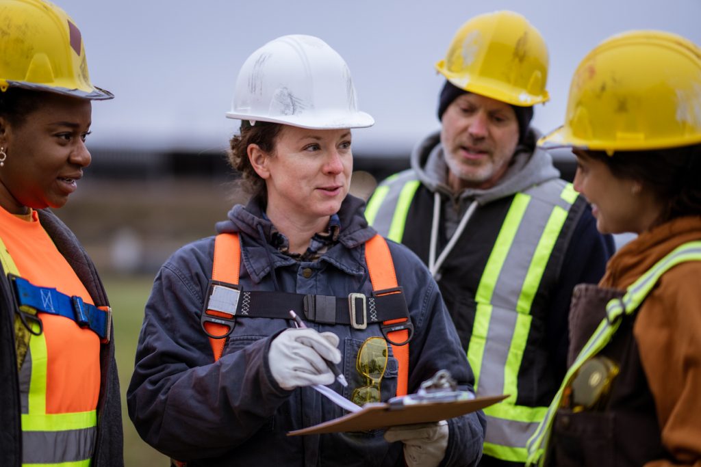 A small group of constructions workers huddle together as they take instructions from the site supervisor. They are each wearing proper protective gear and are listening attentively.